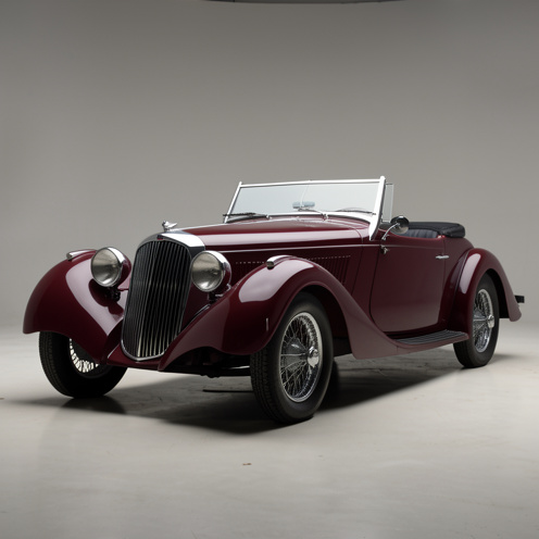 Vue détaillée en trois quarts d’un roadster britannique Jaguar XK 120 OTS de 1953, avec carrosserie bordeaux, calandre chromée, phares ronds et roues à rayons.